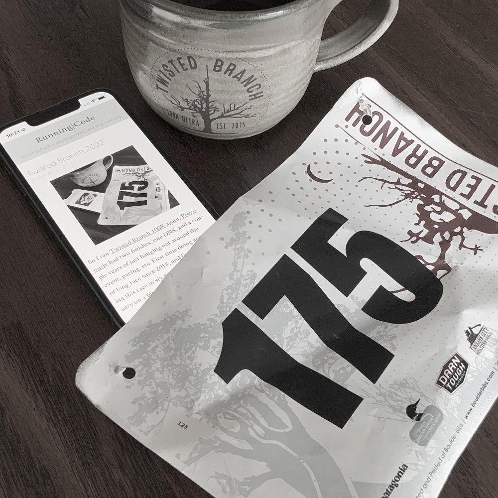 A Twisted Branch finisher's mug full of coffee, my race bib, and a phone showing this blog post.